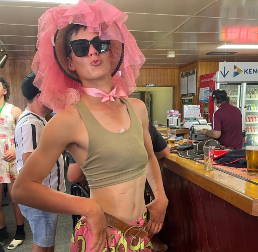 A man stands dressed in a pink frilly bow-peep hat with big sunglasses a brown top and a pink skirt with avocado prints on it