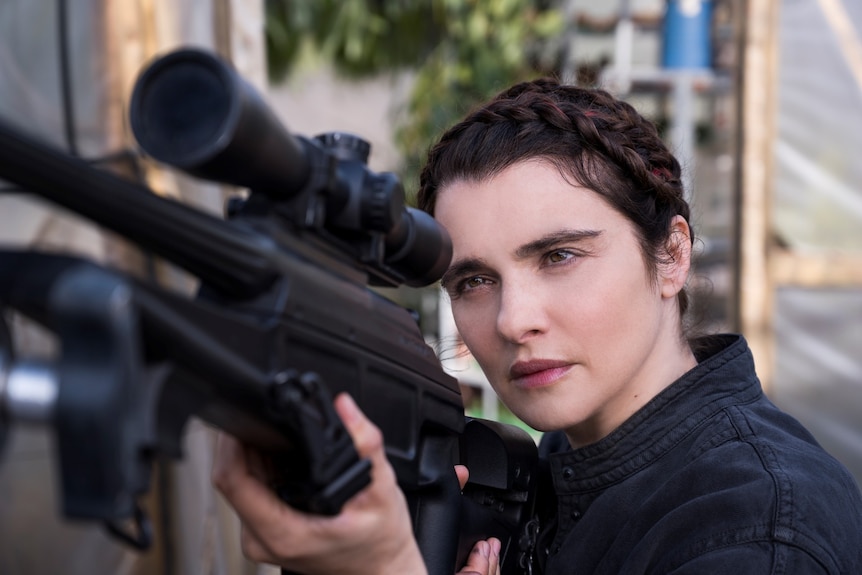 40-something woman with dark hair up in braids, holding automatic rifle on her shoulder and aiming.