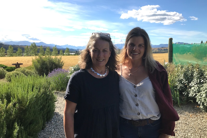 A young woman and her mother stand side by side with green trees and mountains in the background