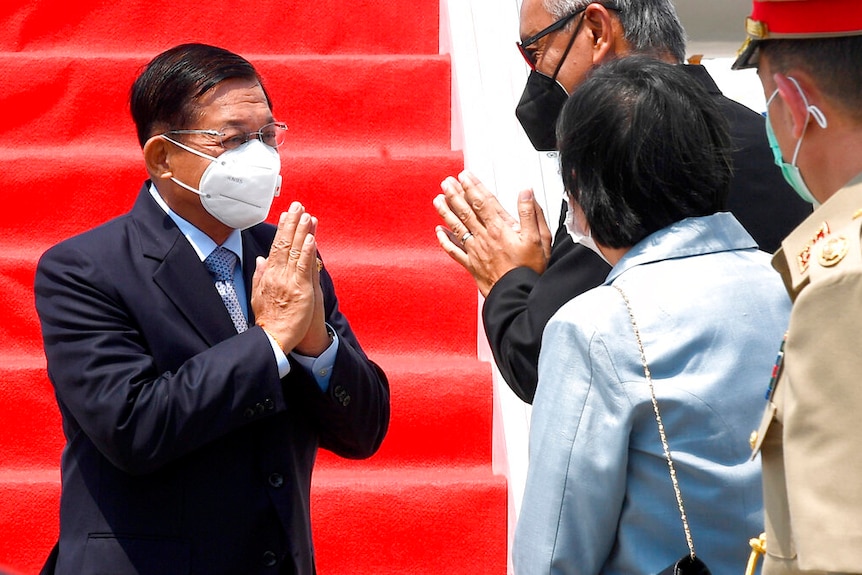 Min Aung Hlaing places his hands together in a greeting, while walking down red carpet laid down on stairs attached to a plane.