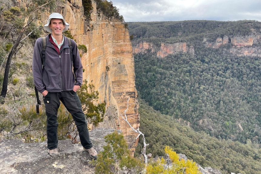 A man with a hat stands on a cliff with a view overlooking some mountains