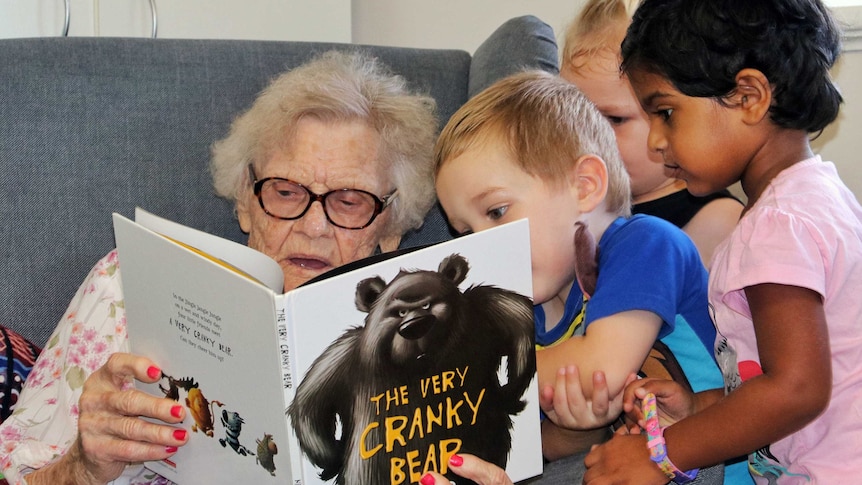 Young children gather around an elderly woman as she reads a book to them.