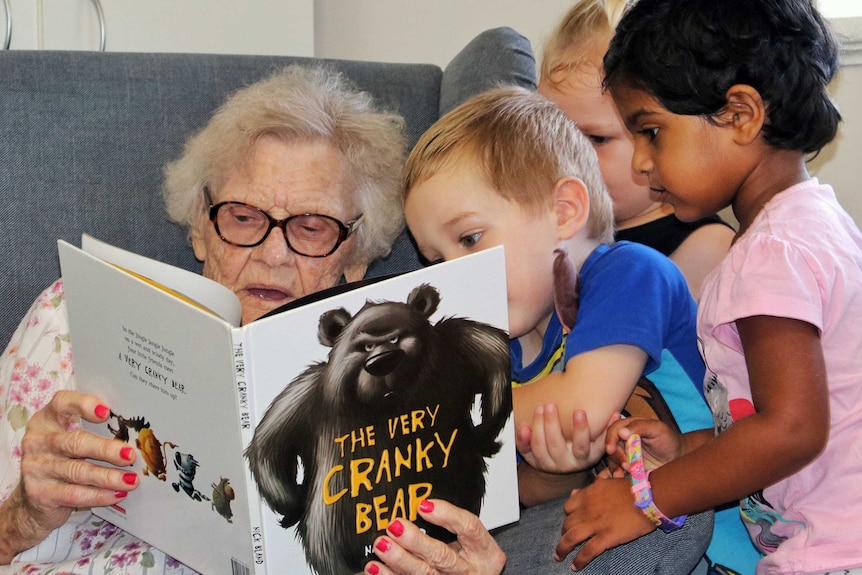 Young children gather around an elderly woman as she reads a book to them.