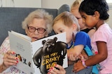 Young children gather around an elderly woman as she reads a book to them.