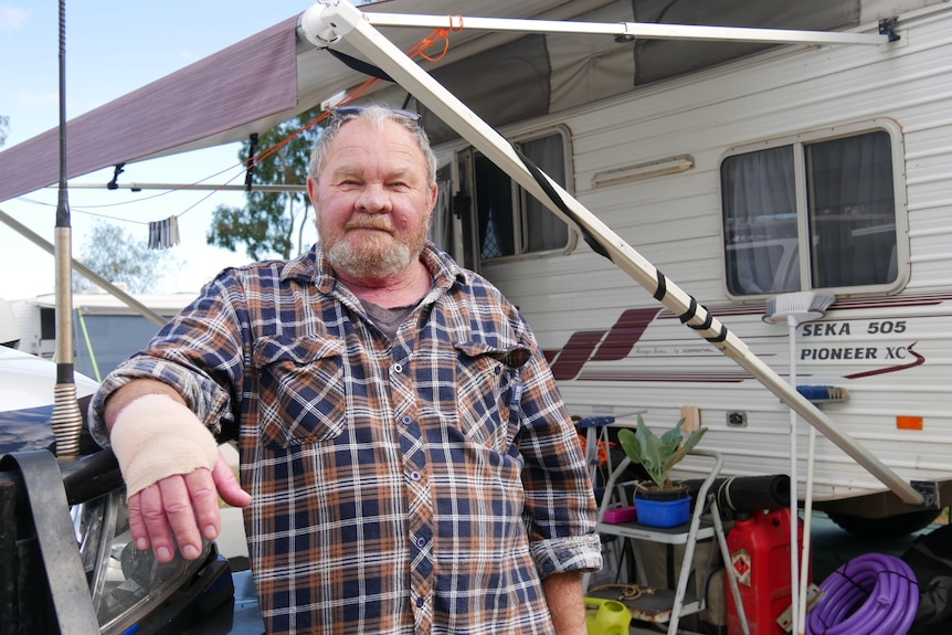 A grey-haired man with a beard and his right arm in a bandage, standing out front of a caravan, wearing a checked shirt