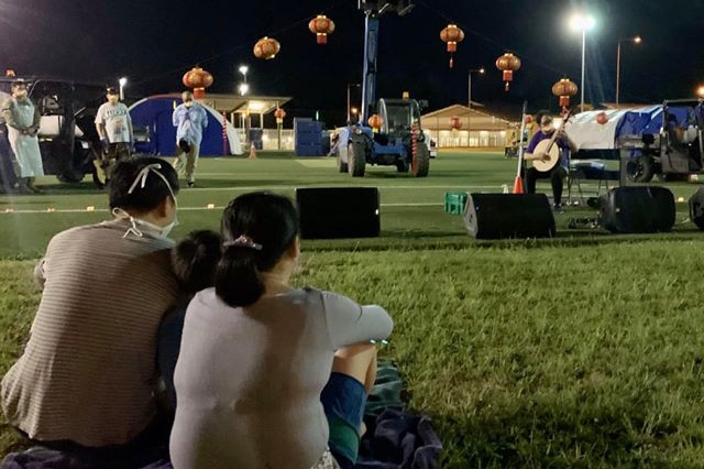 Two people sit on grass at night watching a makeshift concert at Christmas Island Detention Centre, everyone wearing face masks.