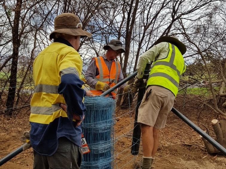 Three men wearing hi vis clothing building a fence with burnt trees in the background