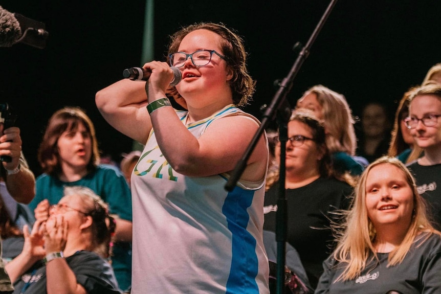 A young woman with down syndrome smiles into a microphone with choir behind