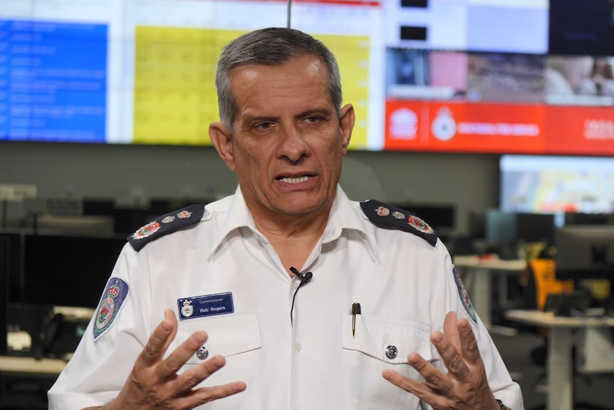 Man in uniform white shirt talking with animated hands with screens in the background 