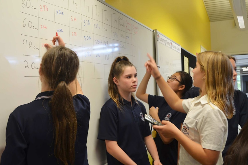 A group of female high school students stand near a whiteboard in a classroom looking at figures written on it.