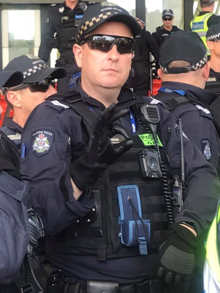 A man in Victoria Police riot gear, wearing black gloves, makes the 'OK' hand gesture