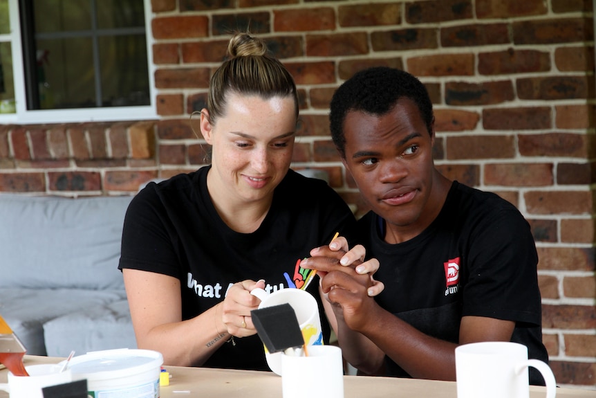 Ashleigh sits at a table with a camp participant and helps him paint a mug.