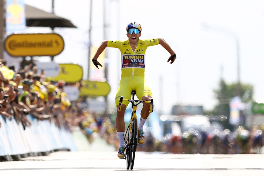 Wout van Aert holds his arms out in celebration while riding over the line wearing the yellow jersey