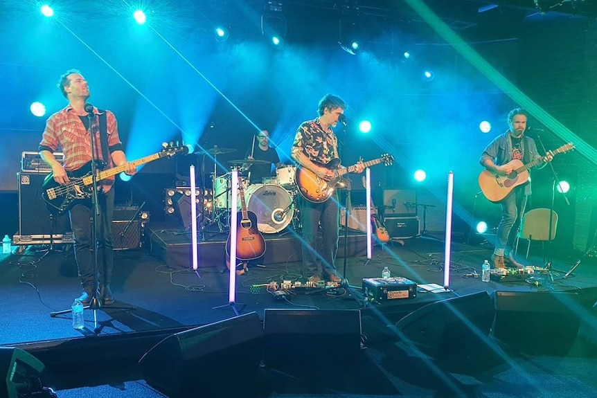 Four members of a band play guitars and drums on a stage