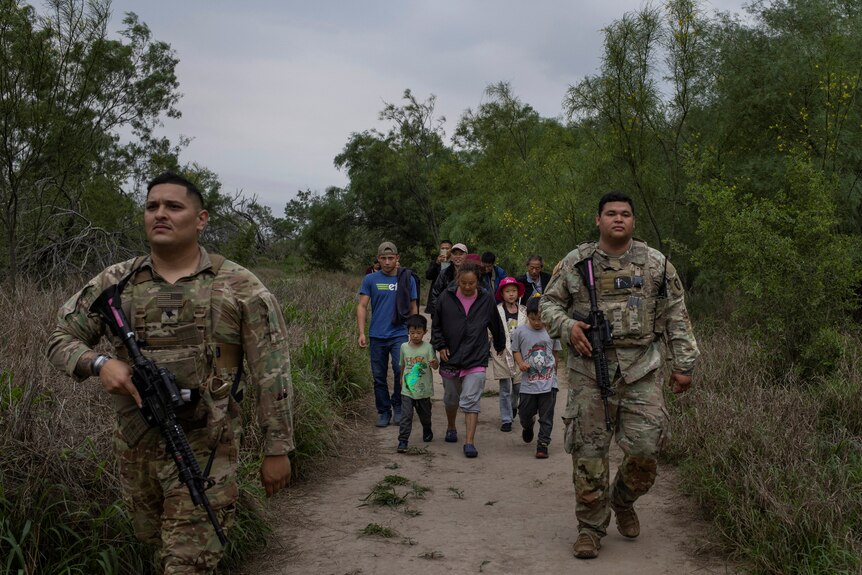 Two men in camouflage carry guns as they walk in front of a group of migrants.
