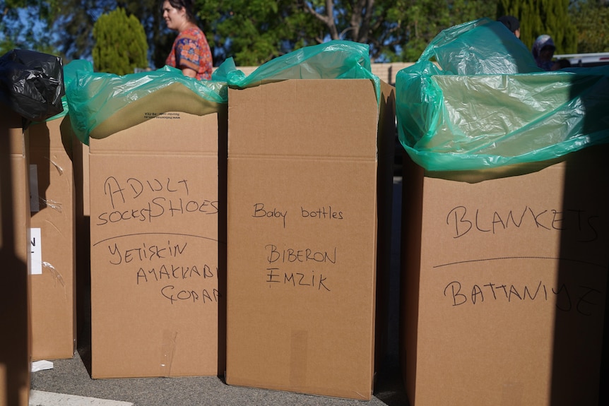 Tall cardboard boxes lined up, labelled in black marker "ADULT shoes", "baby bottles", "blankets".