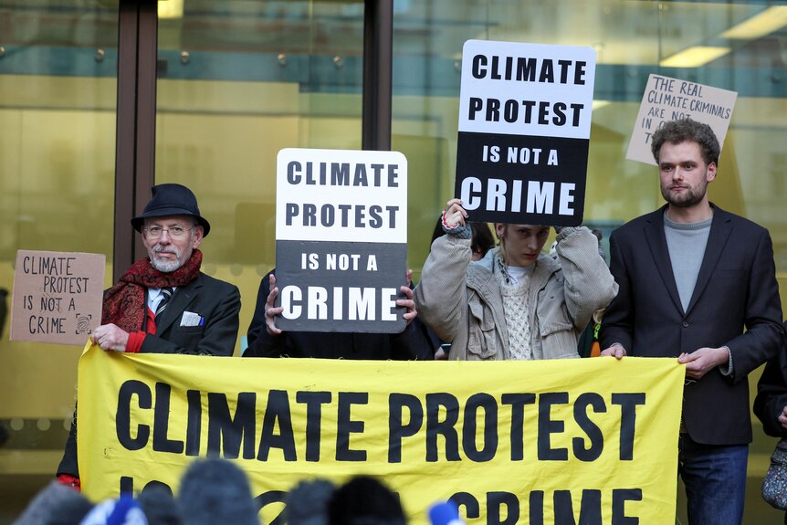 Four people stand behind a yellow banner as two hold signs that say: "Climate protest is not a crime".