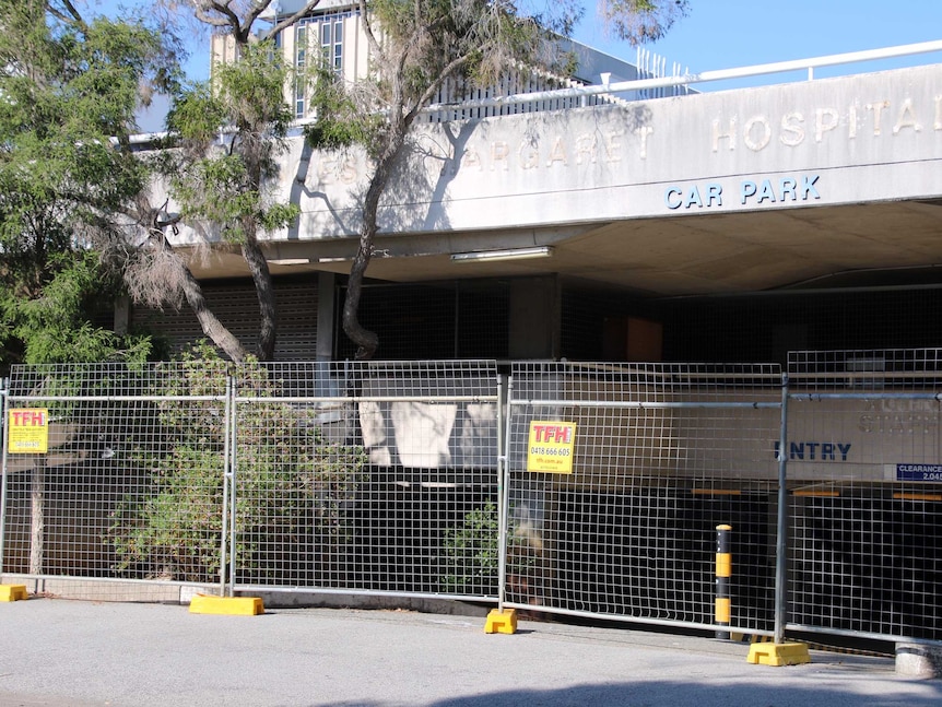 A dilapidated hospital car park sits behind temporary metal fencing.