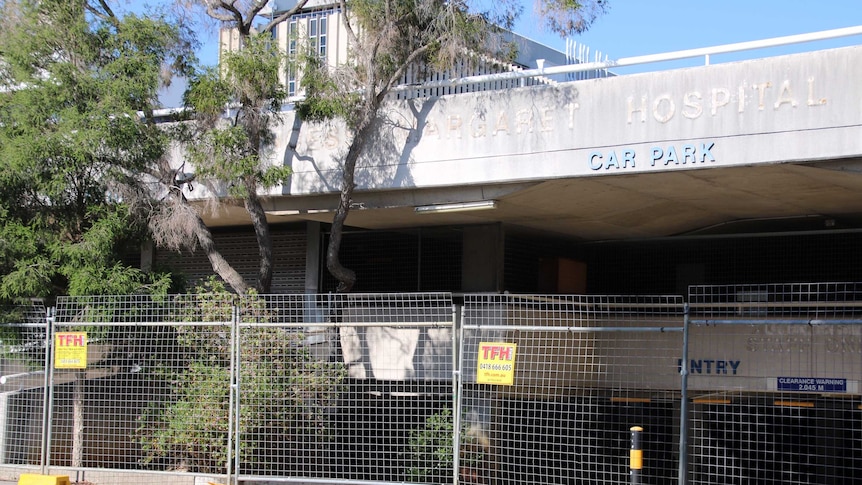 A dilapidated hospital car park sits behind temporary metal fencing.