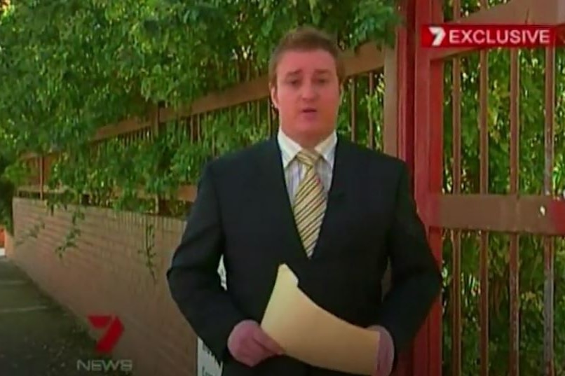 Dylan Howard stands next to a gate and holds a folder while speaking to camera. There's a Channel Seven logo.