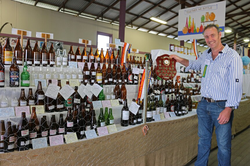 A man stands next to a stand of empty brown, green and clear beer bottles holding a prize shield up.