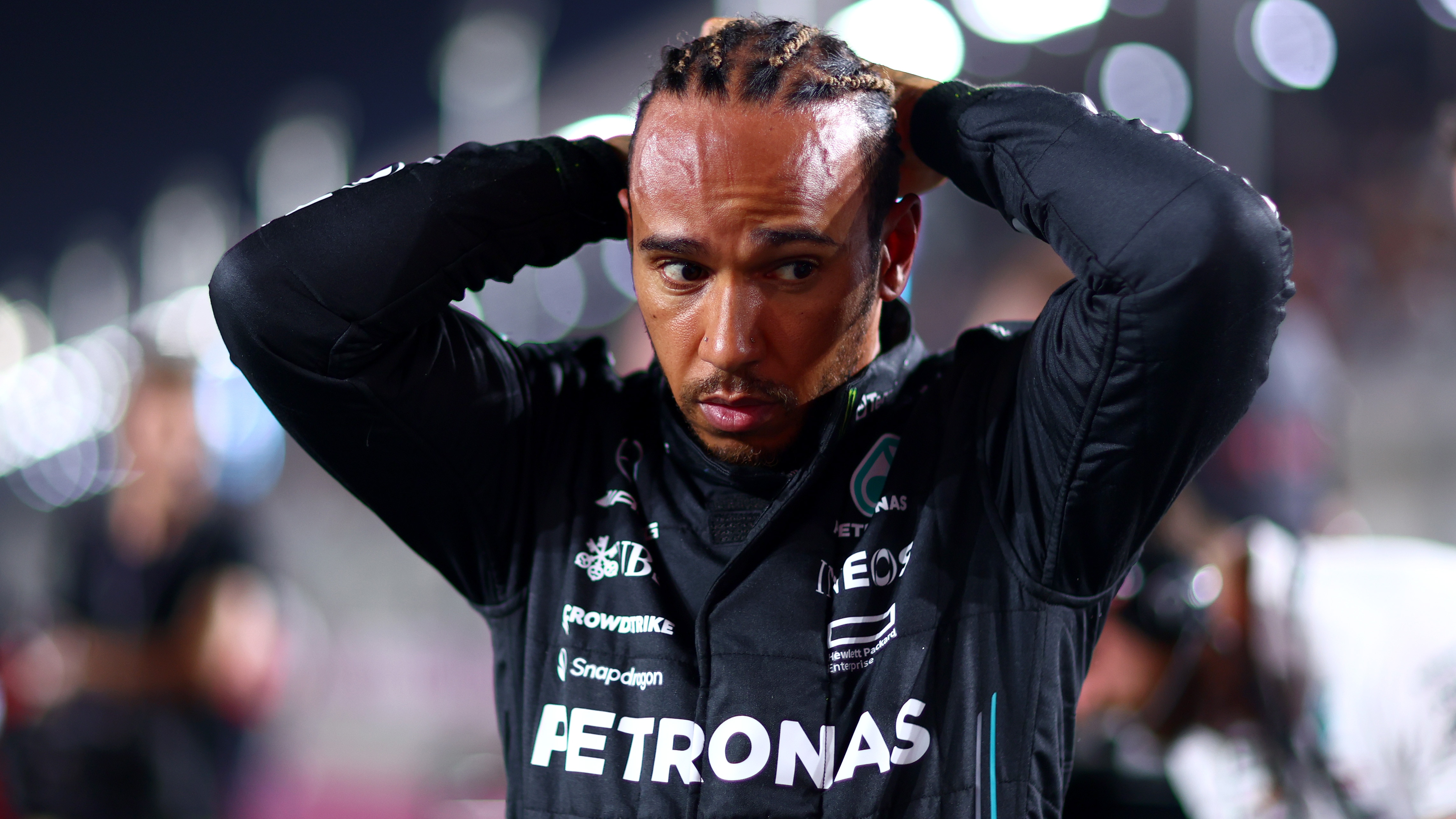 An F1 driver, looking downward, fixing his hair, wearing a black racing suit.