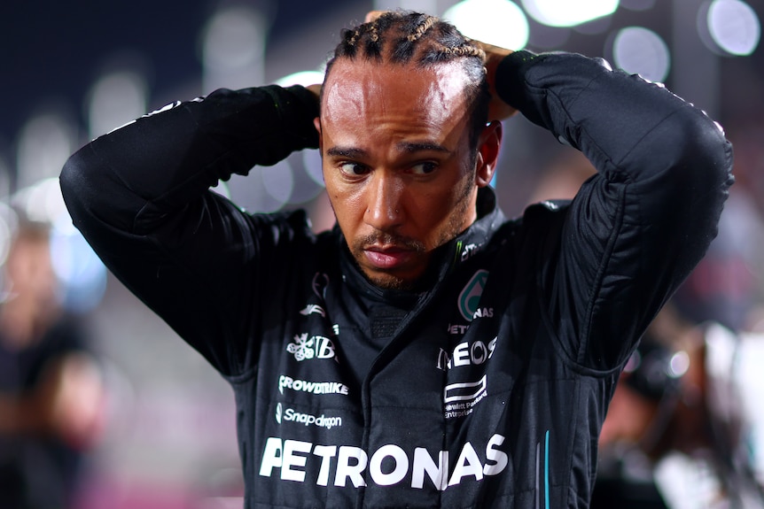 An F1 driver, looking downward, fixing his hair, wearing a black racing suit.