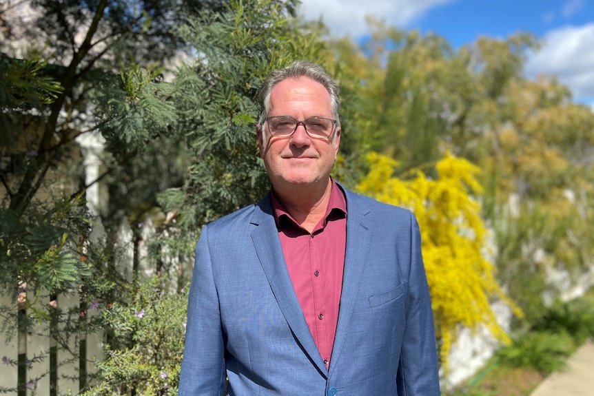 Man standing in front of a bush and wattle, wearing a pink shirt, blue suit jacket and glasses.