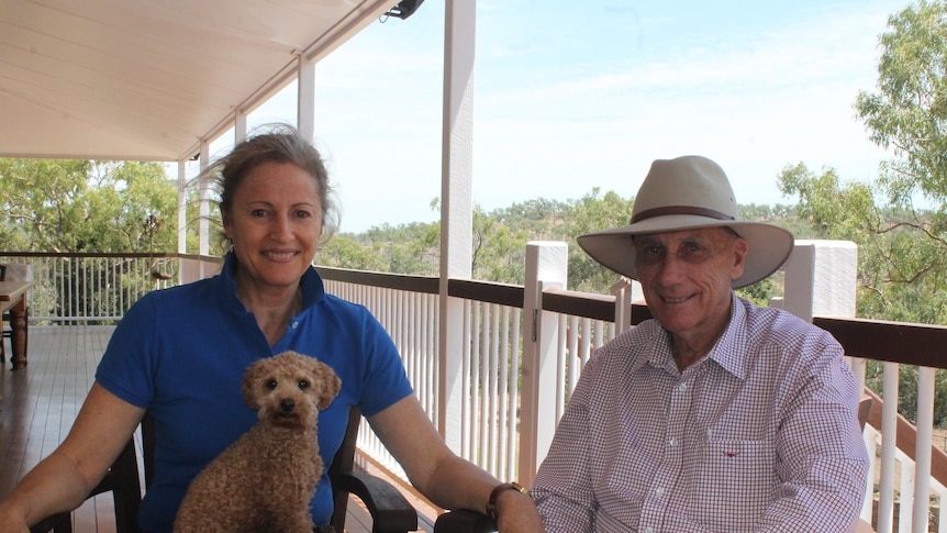 A man and woman sitting together on veranda with their small dog. The man is wearing a large hat.