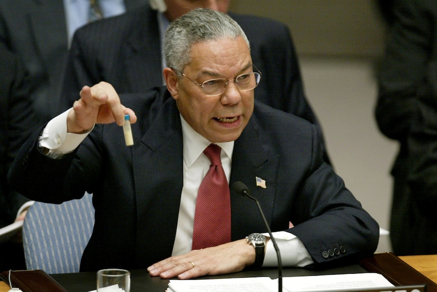 Colin Powell sits at a desk, talking and holding up a small vial in one hand.