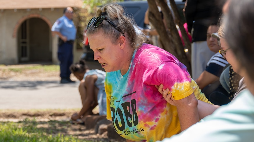 A woman wearing a tie-dye shirt with ROBB Elementary written on the front grimaces in pain as she leans down