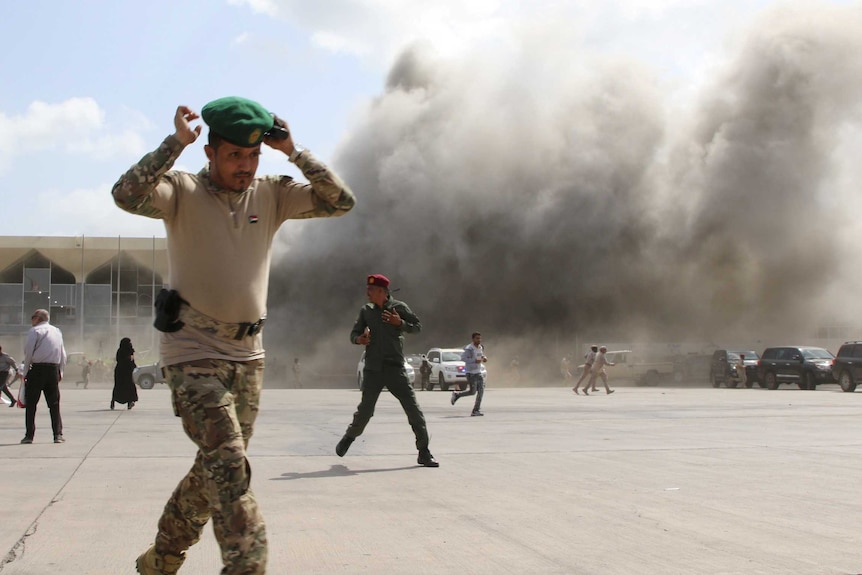 Man in foreground clutches his hat and runs away from blast, several men behind him do the same.