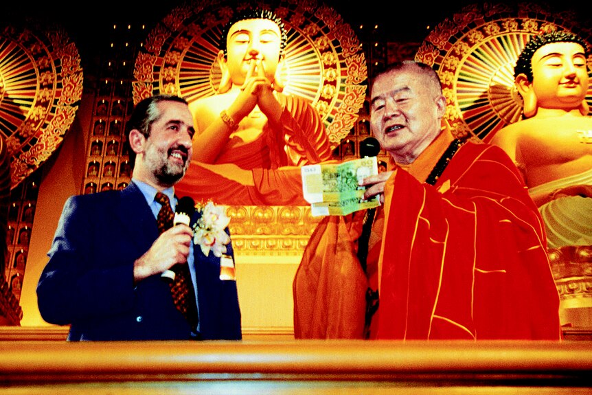 man in suit in front of Buddha statues with a monk holding $100