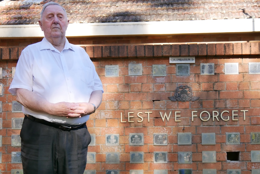 An elderly man in a white button up shirt stands in front of a cremation wall covered in plaques and the words Lest We Forget.