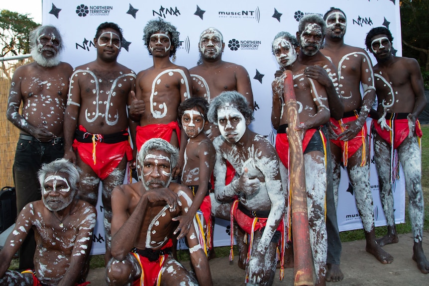 About a dozen Indigenous men of all ages in red loin cloths and white body paint stand in a group with neutral expressions
