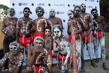 About a dozen Indigenous men of all ages in red loin cloths and white body paint stand in a group with neutral expressions