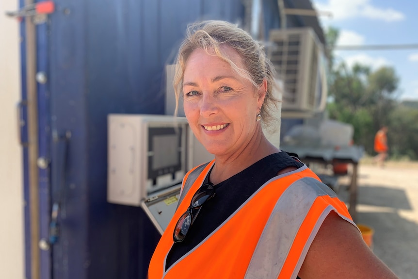 A smiling blonde woman in a high-vis vest stands in front of a shipping container.
