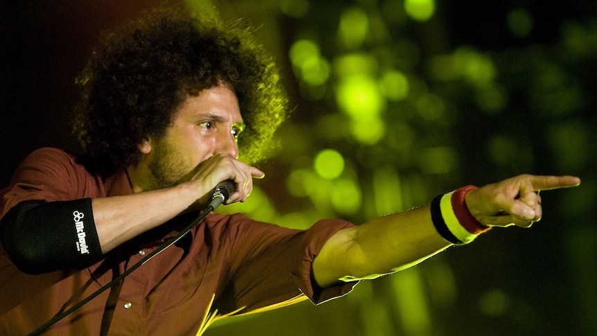 Zach de la Rocha points at the crowd as he raps into a microphone. He wears a brown shirt and armbands and has curly hair.
