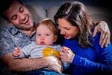 Nas, her husband Tom and their baby Lachie sit closley together at their home. Nas and Tom are laughing. 