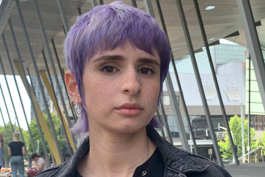 A young woman with short purple hair looks at the camera.