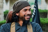 Neil Prakash smiles, glancing to his left. He wears a head scarf and backpack in front a black flag with white writing behind