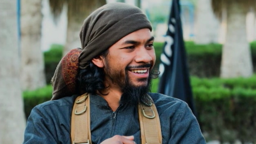 Neil Prakash smiles, glancing to his left. He wears a head scarf and backpack in front a black flag with white writing behind