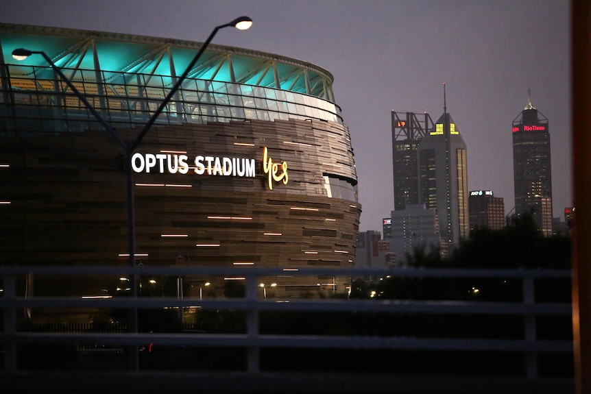A stadium with blue lights at the top and "Optus Stadium Yes" on the side in the evening light in front of the Perth skyline