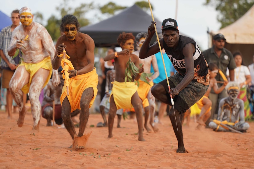 Two dancers, one dressed traditionally, the other wearing western clothes and holding a spear.