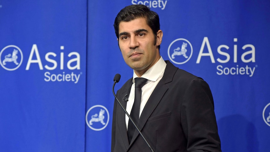 ethnic male in a black and white suit stands behind a microphone and in front of a blue backdrop