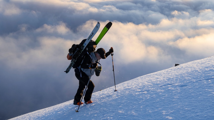 A man in snow gear stands on a snow covered hillside above the clouds with skiis on his back and a hiking pole.