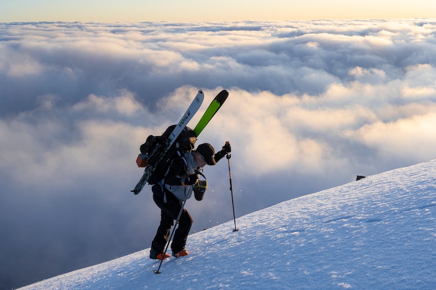 A man in snow gear stands on a snow covered hillside above the clouds with skiis on his back and a hiking pole.