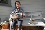 A woman sits on a bench holding some gardens next to a cat wearing a face mask.