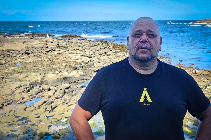 Portrait of man in a t-shirt standing on rocks by the ocean, looking at camera