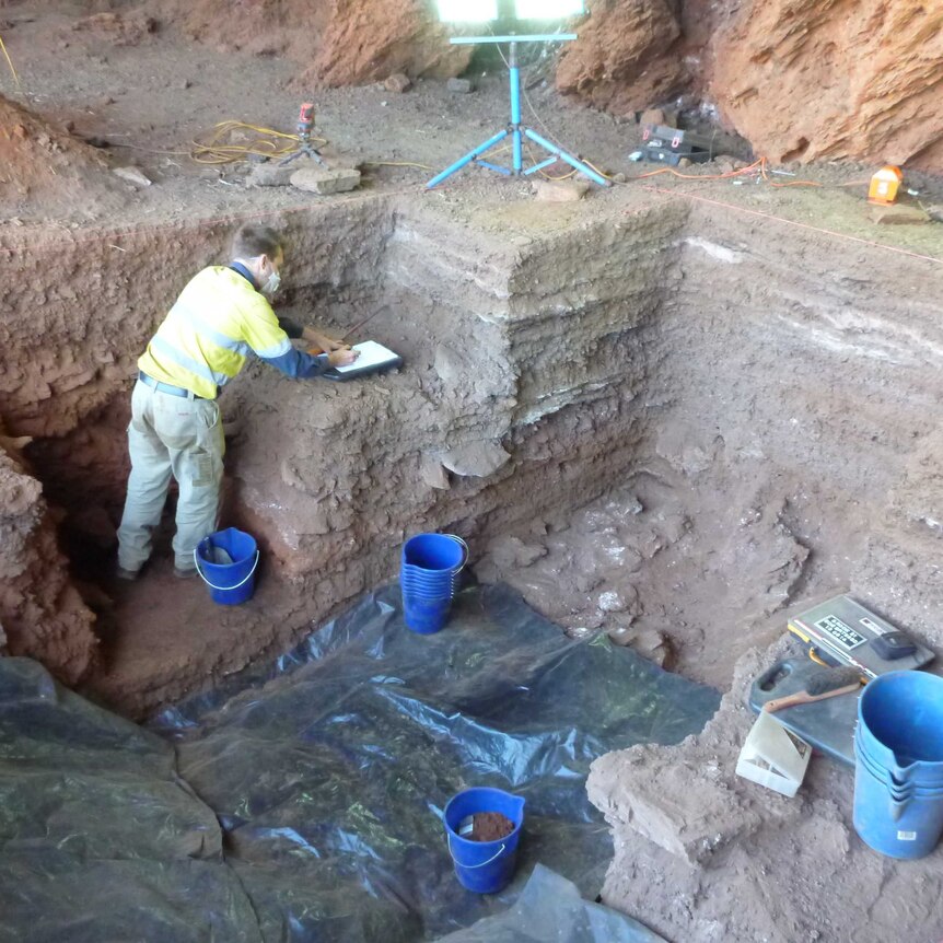 A man inside a dig pit makes notes as he excavates artefacts.